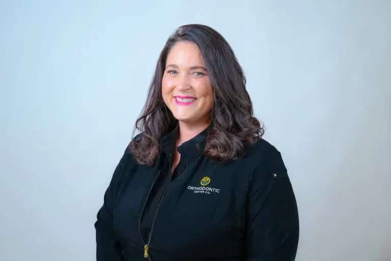 A woman with long dark hair, wearing a black jacket that has the word "ORTHODONTIC" embroidered on it, smiles while standing against a light background.