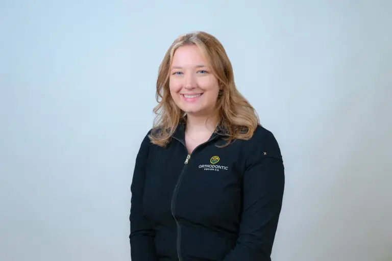 A person with long, light brown hair is smiling and wearing a black jacket with a logo that says "Orthodontic Specialists" against a plain light background.