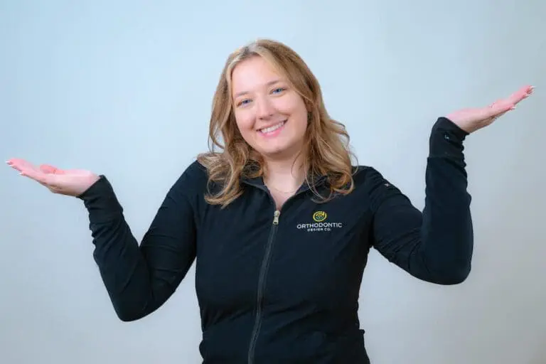 A woman in a black zip-up jacket smiles with both hands raised to the sides against a plain background.