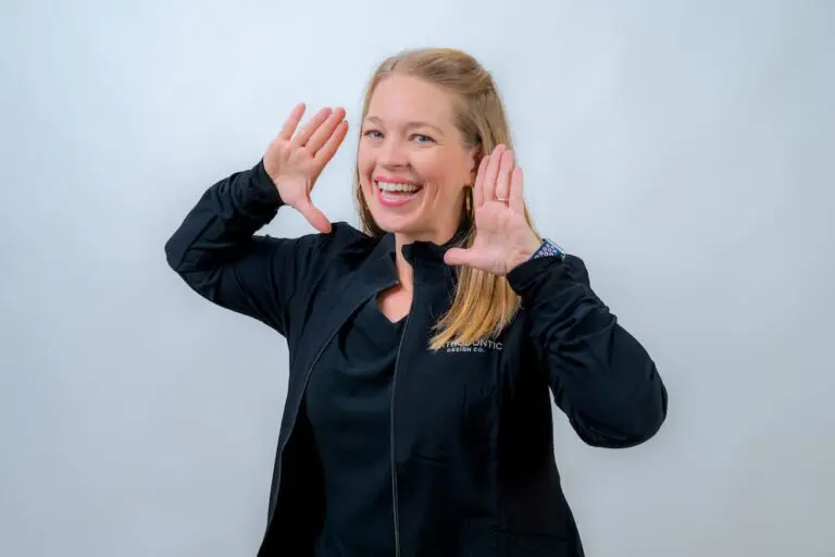 Person with blonde hair in a black jacket smiling and holding hands near face, mimicking a playful gesture against a plain background.