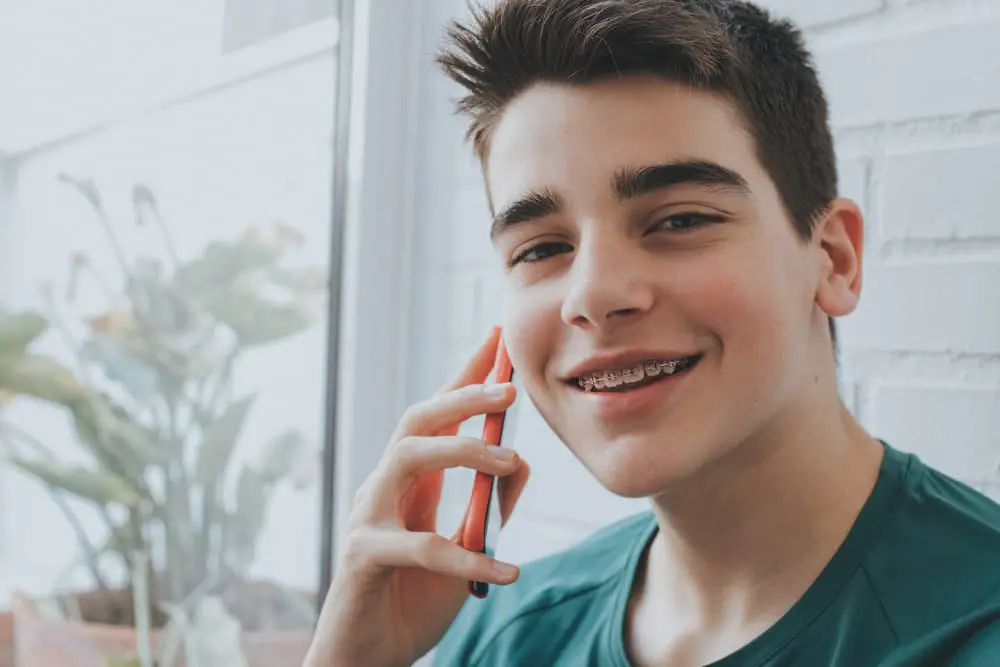 A smiling teenage boy with braces holds a red smartphone to his ear, sitting indoors near a window with plants in the background.