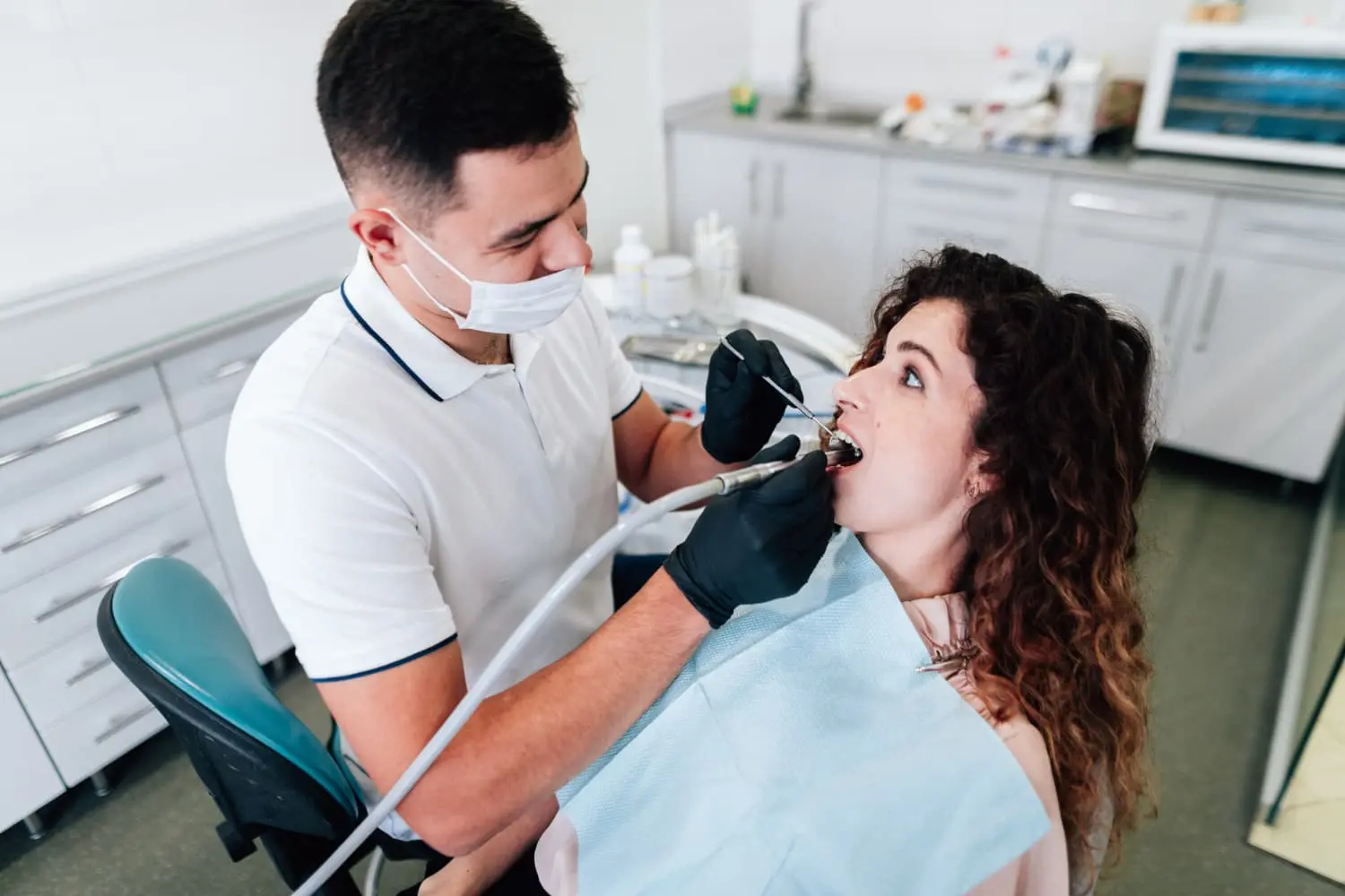 Dentist wearing gloves and a mask examines a woman's teeth using dental tools. The woman is sitting in a dental chair with a blue protective bib.