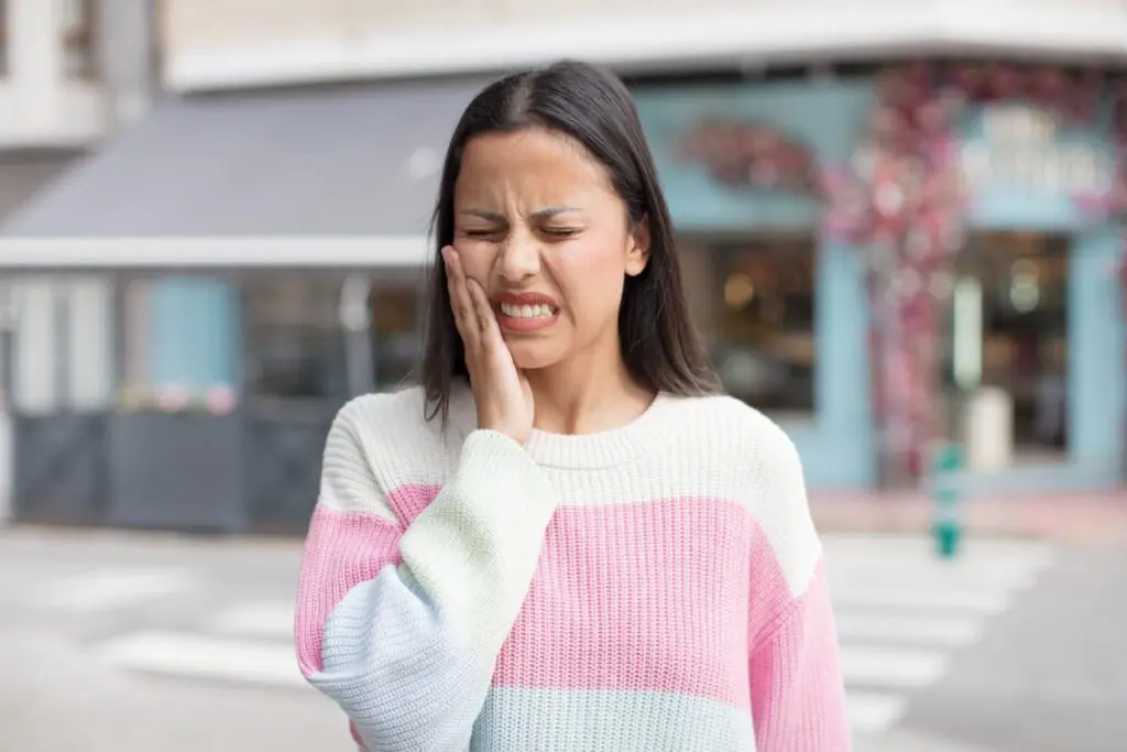 A woman stands outside, touching her cheek with a pained expression, wearing a pastel striped sweater.