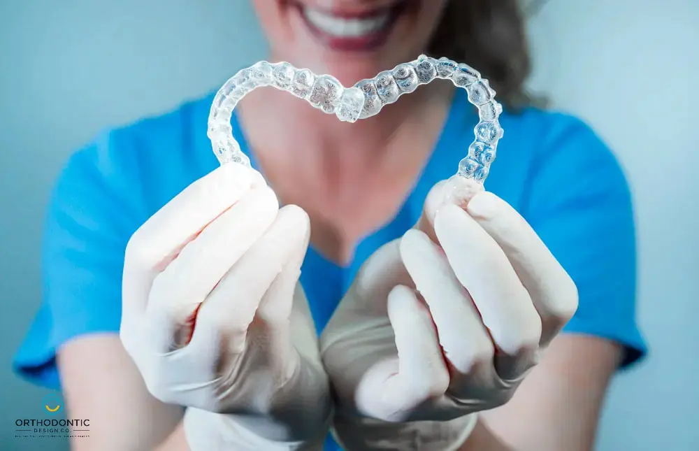 Person in blue scrubs and gloves holds two clear dental aligners forming a heart shape.