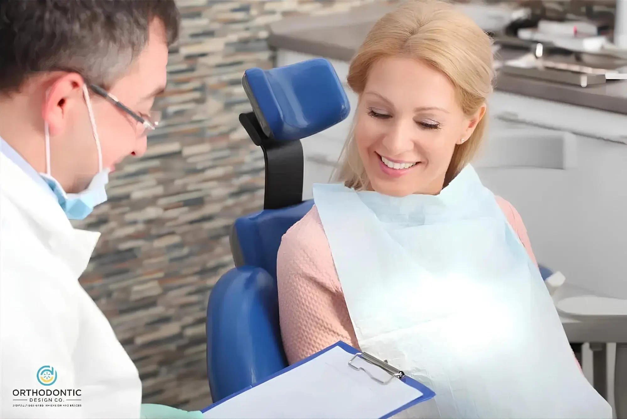 A woman in a dental chair smiles at a dentist holding a clipboard. She is wearing a dental bib and the dentist is wearing a white coat.