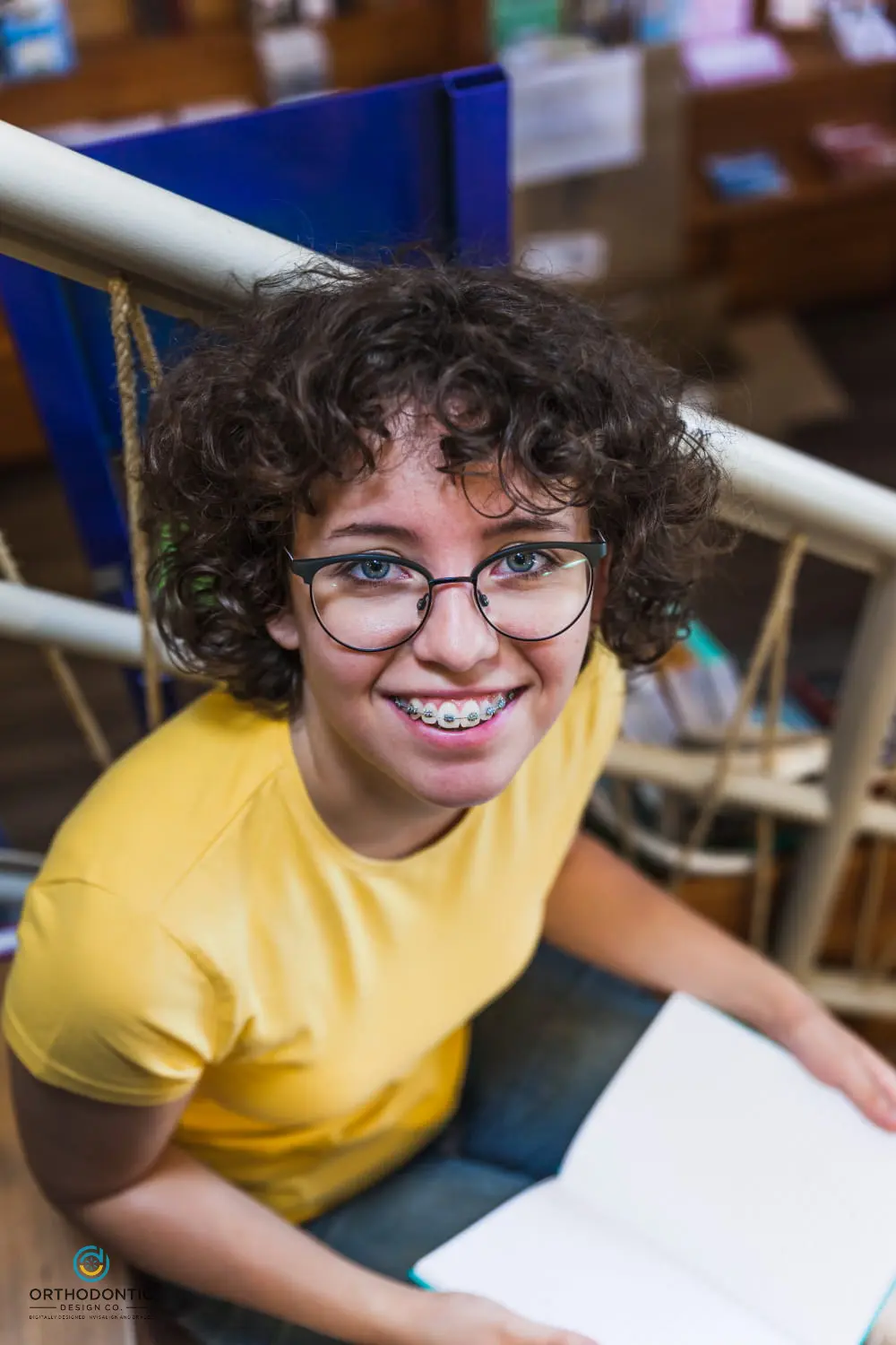 Person with curly hair, wearing glasses and braces, smiles while sitting on stairs and holding an open book.