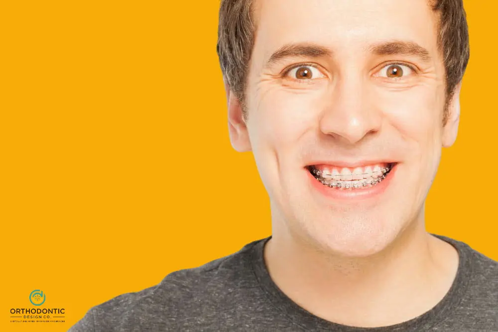 Man smiling with braces against an orange background, wearing a dark shirt. Logo in the bottom left corner.