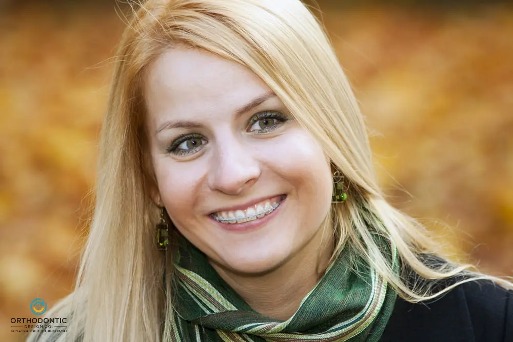 A person with long blonde hair and braces smiles at the camera, wearing a green scarf. Blurred autumn leaves are in the background.