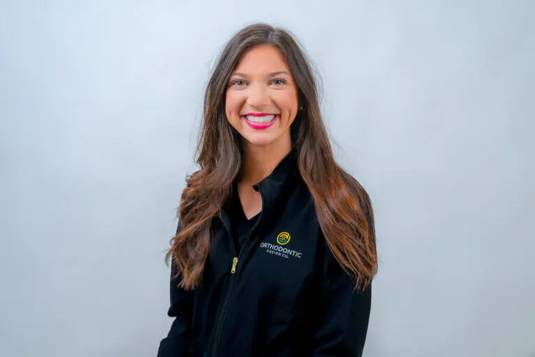 Smiling woman with long hair wearing a black jacket with an orthodontic logo, standing against a plain background.