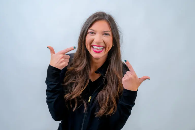 A woman with long brown hair is smiling widely, pointing towards herself with both hands, wearing a black jacket against a light gray background.
