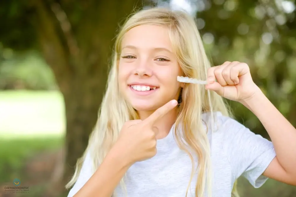 Young girl smiling, holding a clear orthodontic aligner, as one of the alternatives to braces in Madison, AL and pointing at it outdoors.