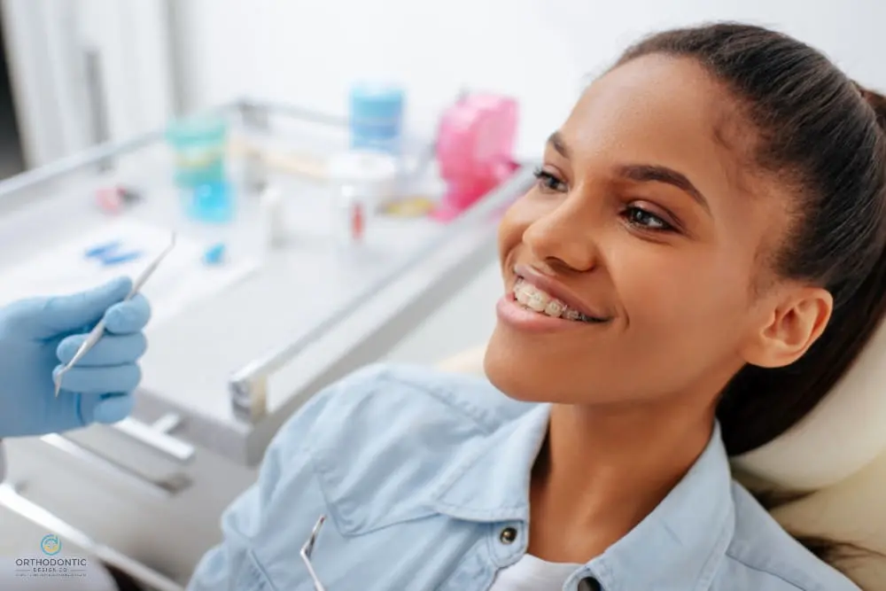 A young woman with clear braces, as one of the alternatives to braces in Madison, AL sits in a dental chair, smiling as a dentist holds a dental tool. Dental equipment is visible in the background.