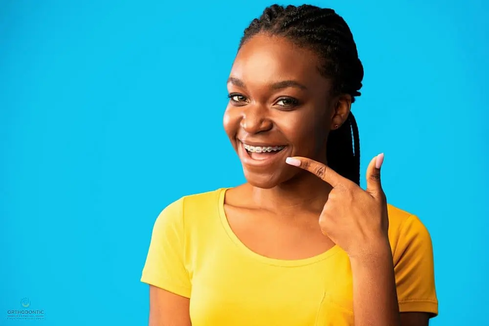 A woman smiles and points to her braces, wearing a yellow shirt against a blue background.