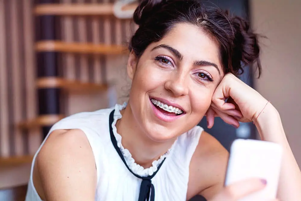 A woman with braces smiles while holding a smartphone. She is wearing a white sleeveless top and has her hair in two buns in Huntsville, AL