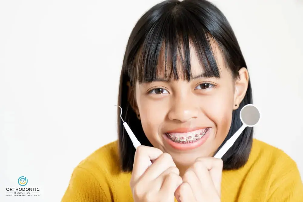 A young person with metal braces smiling and holding dental tools in both hands at Orthodontic Design Co in Huntsville, AL