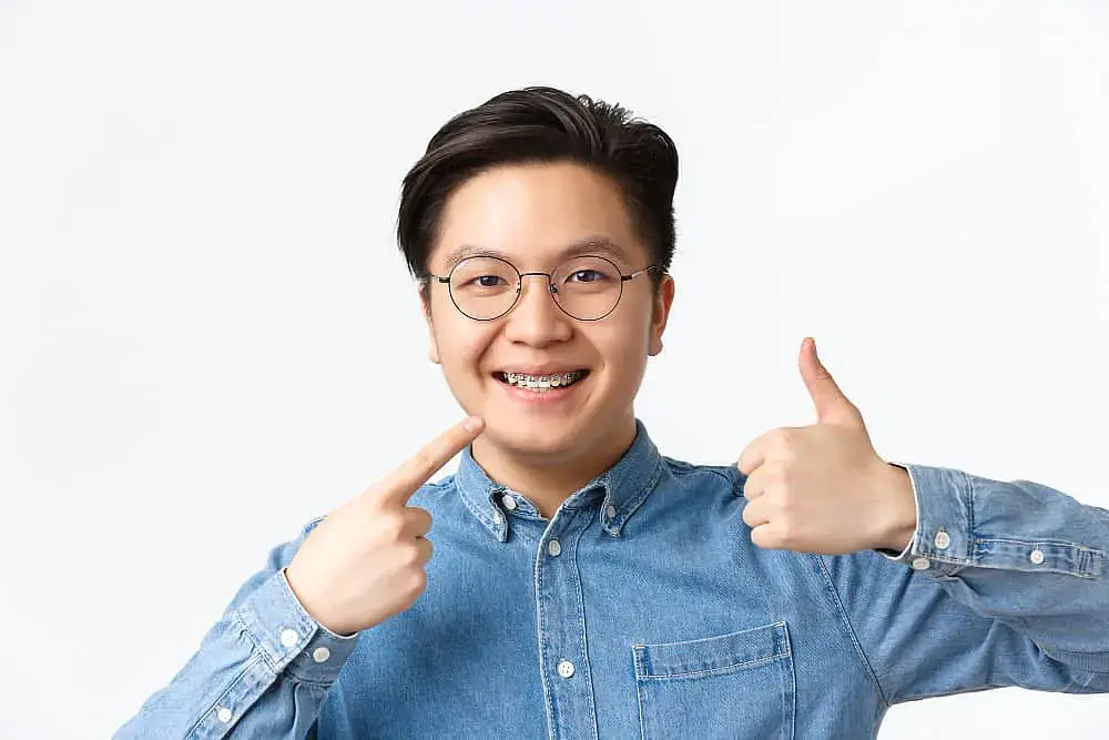 A person wearing round glasses and metal braces smiles, pointing to their braces with one hand and giving a thumbs-up with the other. They are wearing a denim shirt against a plain background in Madison and Huntsville, AL A person wearing round glasses and metal braces smiles, pointing to their braces with one hand and giving a thumbs-up with the other. They are wearing a denim shirt against a plain background in Madison and Huntsville, AL
