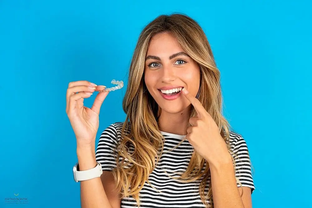 A woman in a striped shirt holds a dental aligner, smiling and pointing at her teeth, with a blue background after considering Aligners vs Braces in Madison, AL.