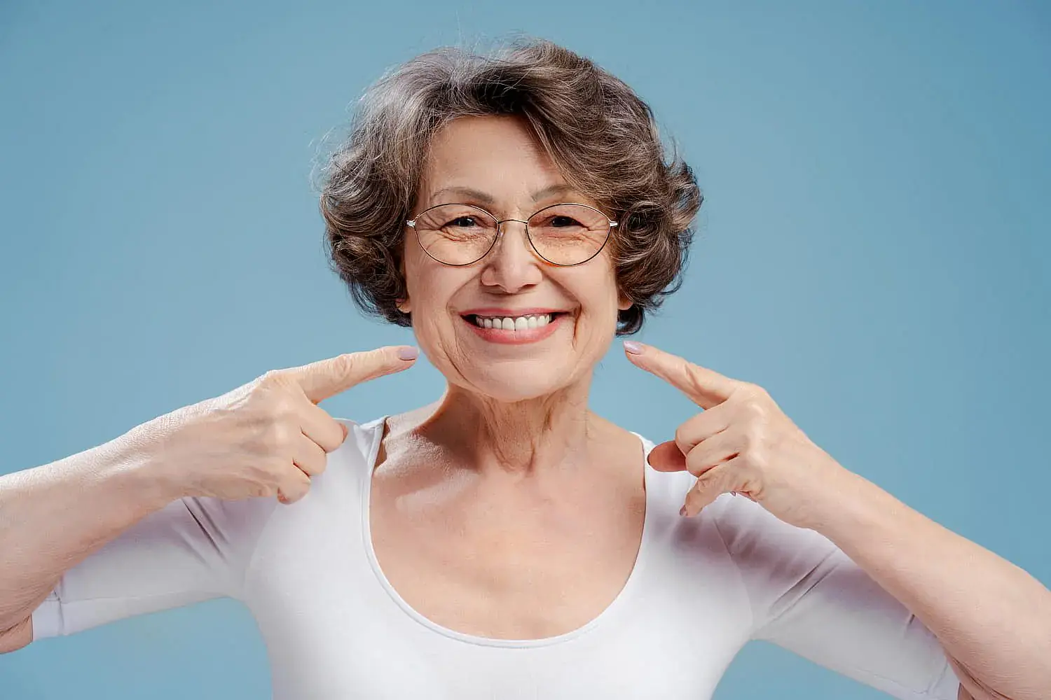 An older woman with glasses smiles confidently and points to her teeth against a blue background, showcasing the results possible with Orthodontic Design Company in Madison and Huntsville, AL—where orthodontic insurance plans support your smile. An older woman with glasses smiles confidently and points to her teeth against a blue background, showcasing the results possible with Orthodontic Design Company in Madison and Huntsville, AL—where orthodontic insurance plans support your smile.