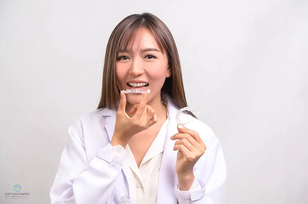 A person in a white coat smiles at the camera while inserting a clear dental aligner—an approach offered by Orthodontic Design Co. Orthodontics for how to fix overbite in Madison and Huntsville, AL