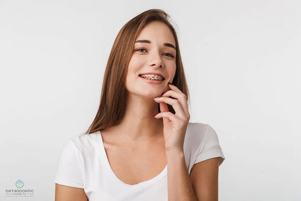 A young woman with long brown hair, wearing a white shirt, smiles and shows her metal braces—Orthodontic Design Co. Orthodontics in Madison and Huntsville, AL—posing against a plain white background after understanding about how long do braces take in madison and huntsville, al.