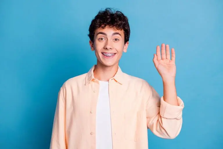 A smiling teenager with curly hair and braces, dressed in a light peach shirt over a white t-shirt, raises their right hand against a blue background—curious about “How Long Do Braces Take in Madison and Huntsville, AL."