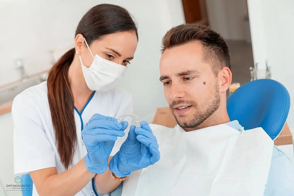 At Orthodontic Design Co. Orthodontics in Madison and Huntsville, AL, a dentist wearing gloves and a mask demonstrates a clear dental aligner to a seated male patient and does insurance cover invisalign in madison and huntsville, al.