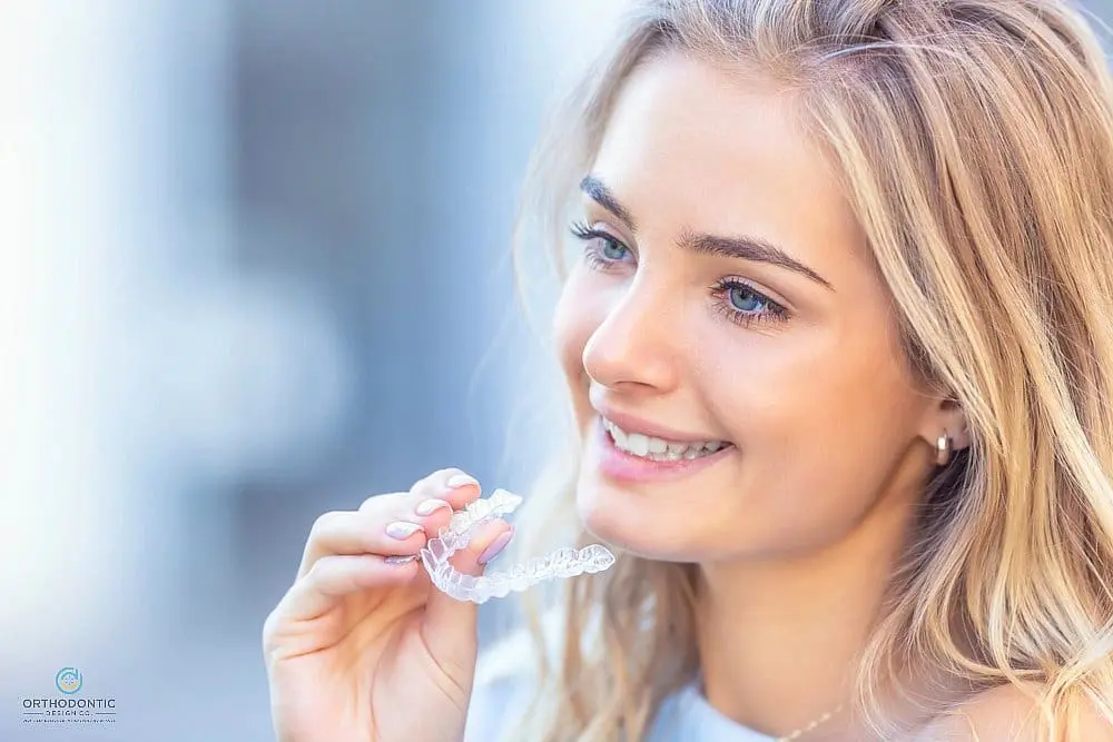 A young woman smiles as she prepares to insert a clear aligner, showcasing does insurance cover invisalign in madison and huntsville, al and the advanced care at Orthodontic Design Co. Orthodontics in Madison and Huntsville, AL.