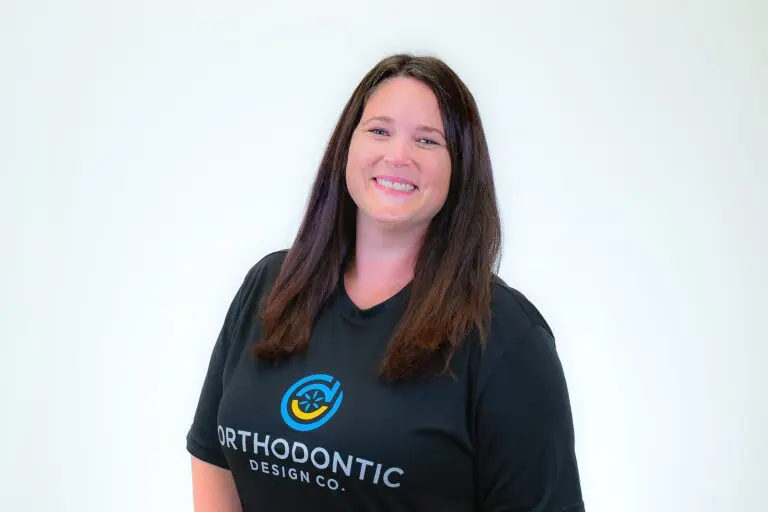 A woman with long brown hair, wearing a black "Orthodontic Design Co. Orthodontics" t-shirt, smiles against a plain white background in Madison and Huntsville, AL.