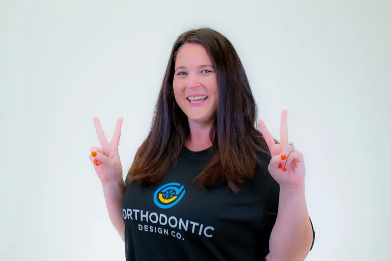 A woman with long brown hair smiles at the camera, wearing a black "Orthodontic Design Co." t-shirt and flashing peace signs, representing Orthodontic Design Co. Orthodontics in Madison and Huntsville, AL.