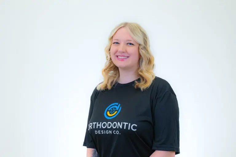 A smiling blonde woman in a black Orthodontic Design Co. Orthodontics t-shirt stands against a plain white background, representing the practice in Madison and Huntsville, AL.