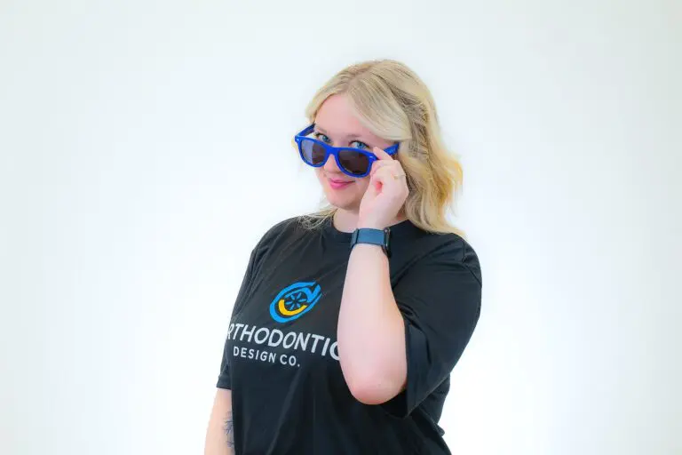 A person with blonde hair, wearing blue sunglasses and a black "Orthodontic Design Co." t-shirt, looks at the camera against a white background, representing Orthodontic Design Co. Orthodontics in Madison and Huntsville, AL.
