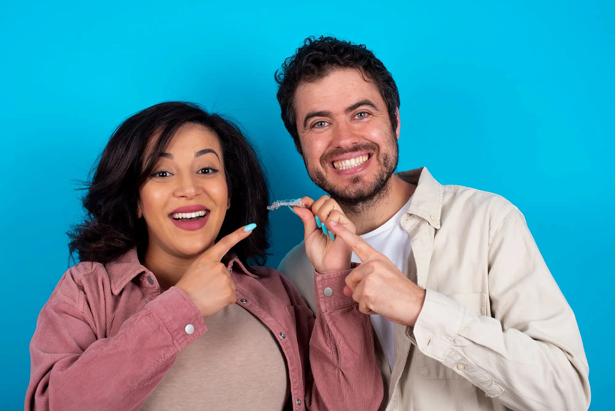A man and woman smile against a blue background, pointing at an Invisalign aligner used by the best orthodontist in Huntsville, AL.