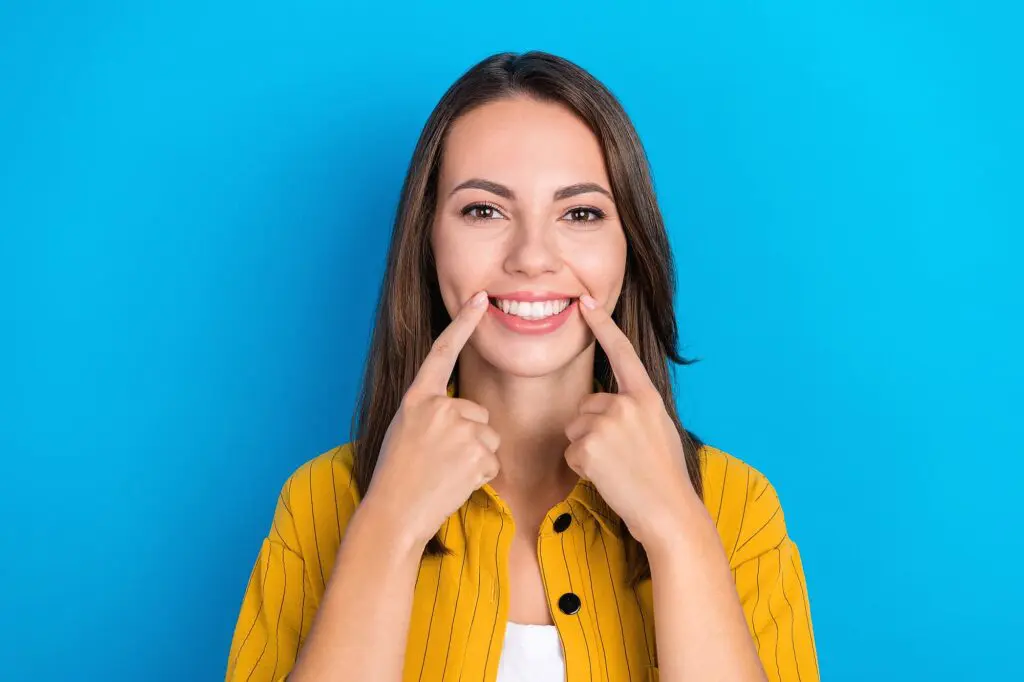 A woman in a yellow shirt stands against a blue background, smiling and pointing to her mouth with both index fingers—highlighting the confident results from Orthodontic Design Co. Orthodontics in Madison and Huntsville, AL with lingual braces or braces behind youth teeth