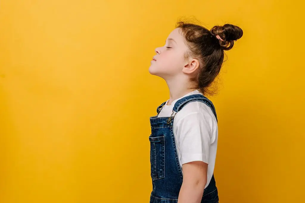 A young girl wearing denim overalls and a white shirt stands in profile with her eyes closed showing proper breathing and airway for good growth in orhtodontics