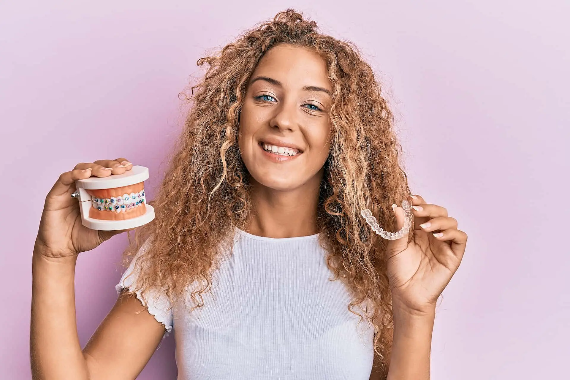 A woman with curly hair stands against a pink background, holding both a dental model with braces and a clear aligner to highlight adult orthodontics options available at Orthodontic Design Co. Orthodontics in Madison and Huntsville, AL.