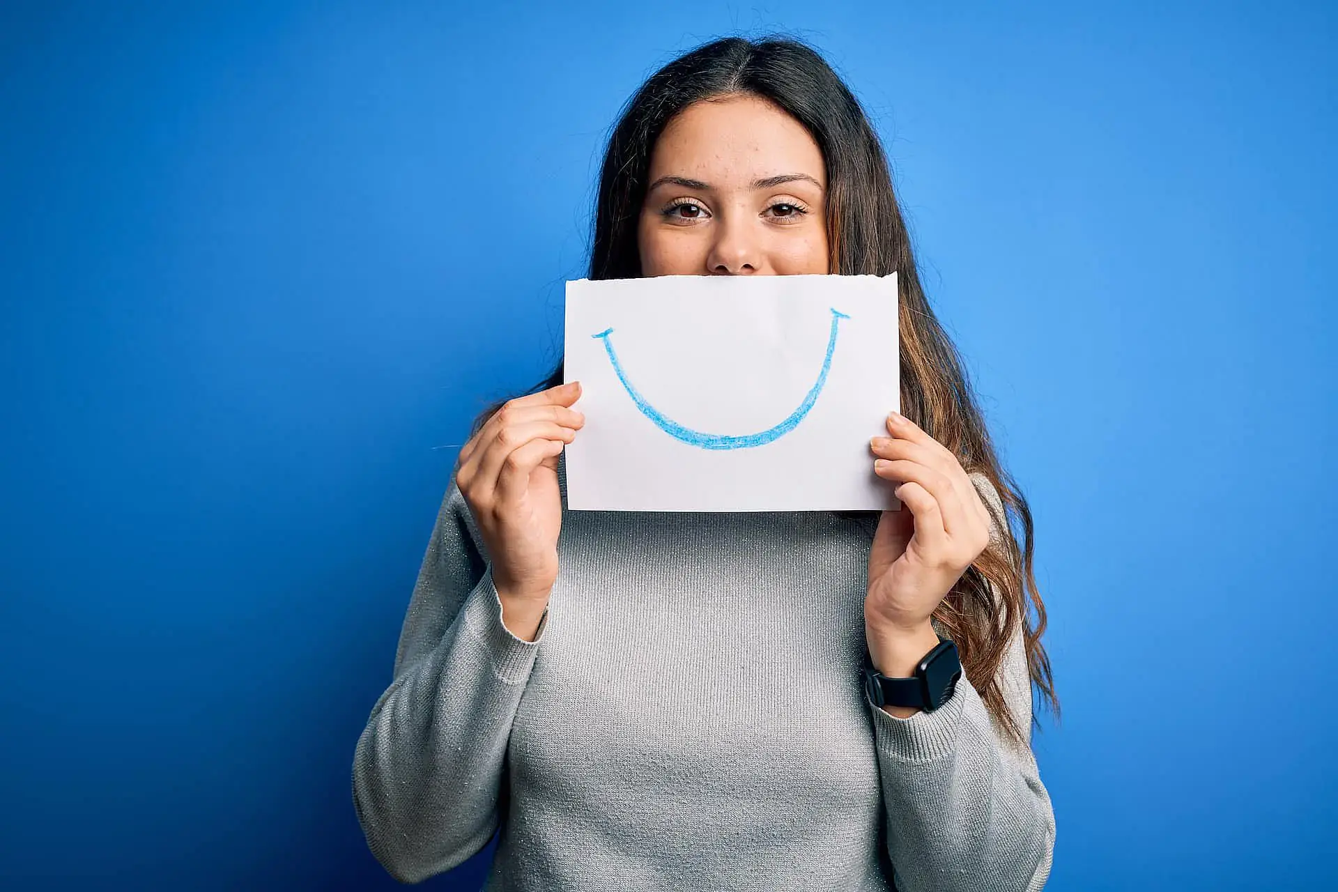 A woman stands against a blue background, playfully holding a paper with a large blue smile in front of her mouth—reflecting the confidence gained through adult orthodontics from Orthodontic Design Co. Orthodontics in Madison and Huntsville, AL. A woman stands against a blue background, playfully holding a paper with a large blue smile in front of her mouth—reflecting the confidence gained through adult orthodontics from Orthodontic Design Co. Orthodontics in Madison and Huntsville, AL.