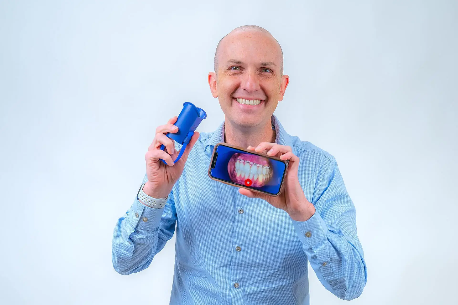 Dr. Adam Reynolds, a children’s orthodontist at Orthodontic Design Co. Orthodontics in Madison and Huntsville, AL, smiles at the camera while holding a blue case and a smartphone showing a dental image with a red light. Dr. Adam Reynolds, a children’s orthodontist at Orthodontic Design Co. Orthodontics in Madison and Huntsville, AL, smiles at the camera while holding a blue case and a smartphone showing a dental image with a red light.