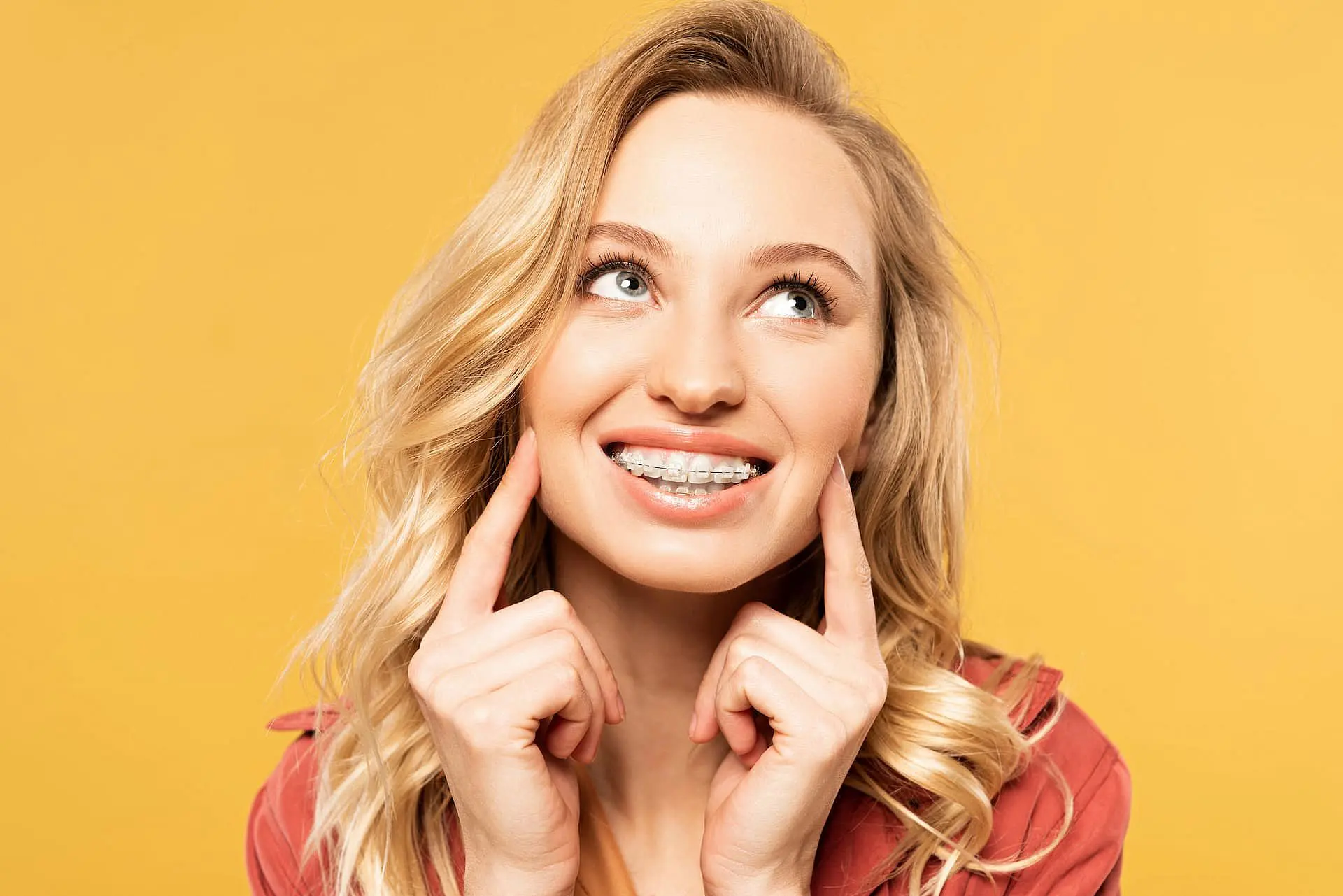 A young blonde woman smiles and points to her braces, highlighting the benefits of adult orthodontics provided by Orthodontic Design Co. Orthodontics in Madison and Huntsville, AL, as she stands against a yellow background.