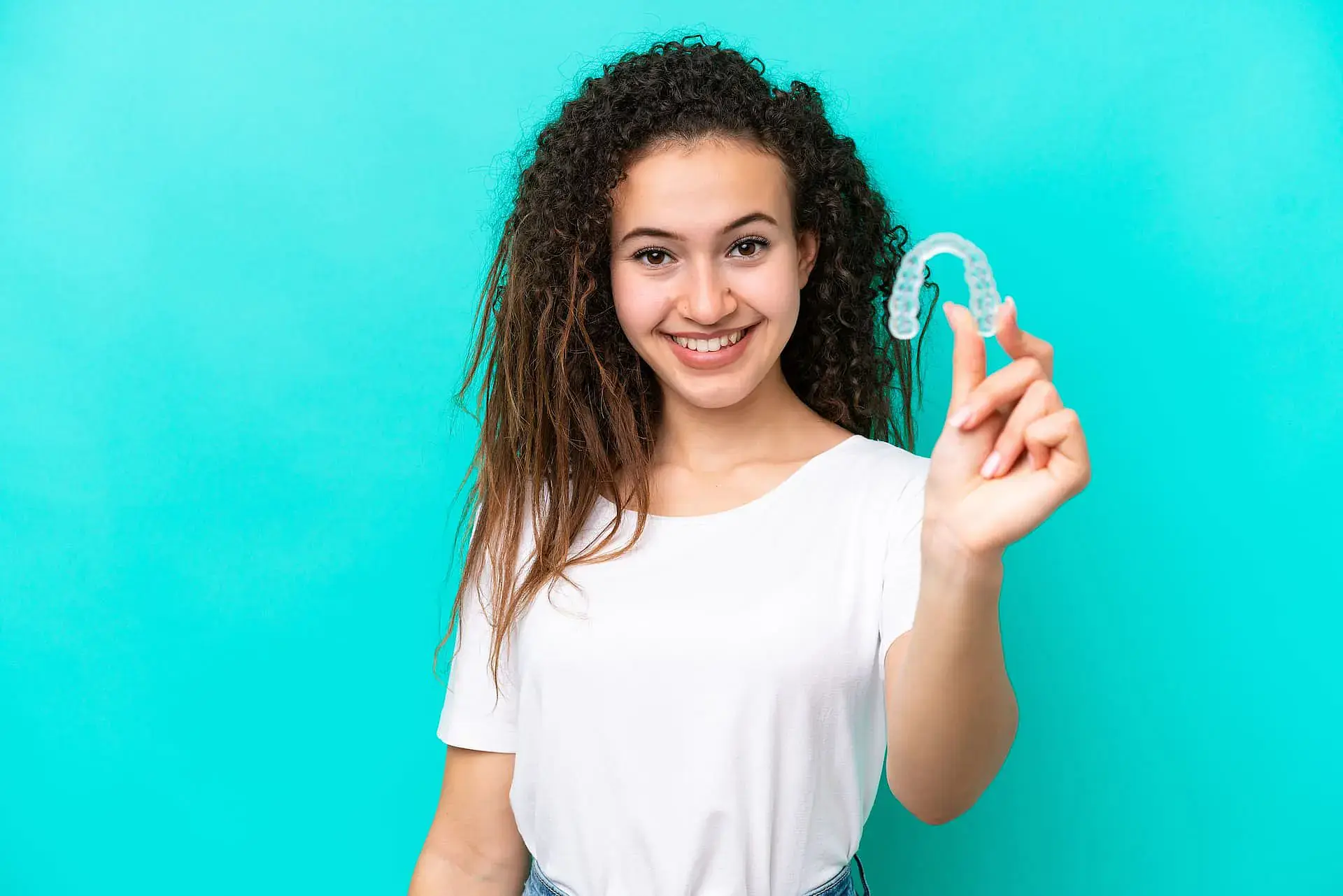 A young woman in a white shirt smiles and holds a clear dental aligner—often recommended as retainers by children’s orthodontists—for Orthodontic Design Co. Orthodontics in Madison and Huntsville, AL, set against a turquoise background. A young woman in a white shirt smiles and holds a clear dental aligner—often recommended as retainers by children’s orthodontists—for Orthodontic Design Co. Orthodontics in Madison and Huntsville, AL, set against a turquoise background.