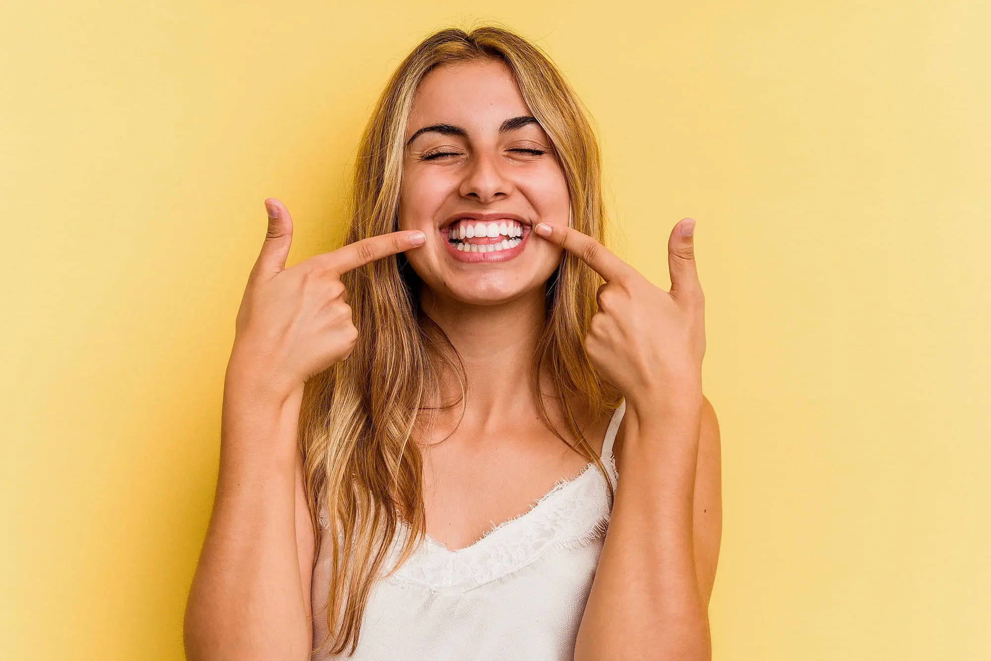 A young woman with long blond hair smiles and points to her teeth with hidden braces in Huntsville, AL.