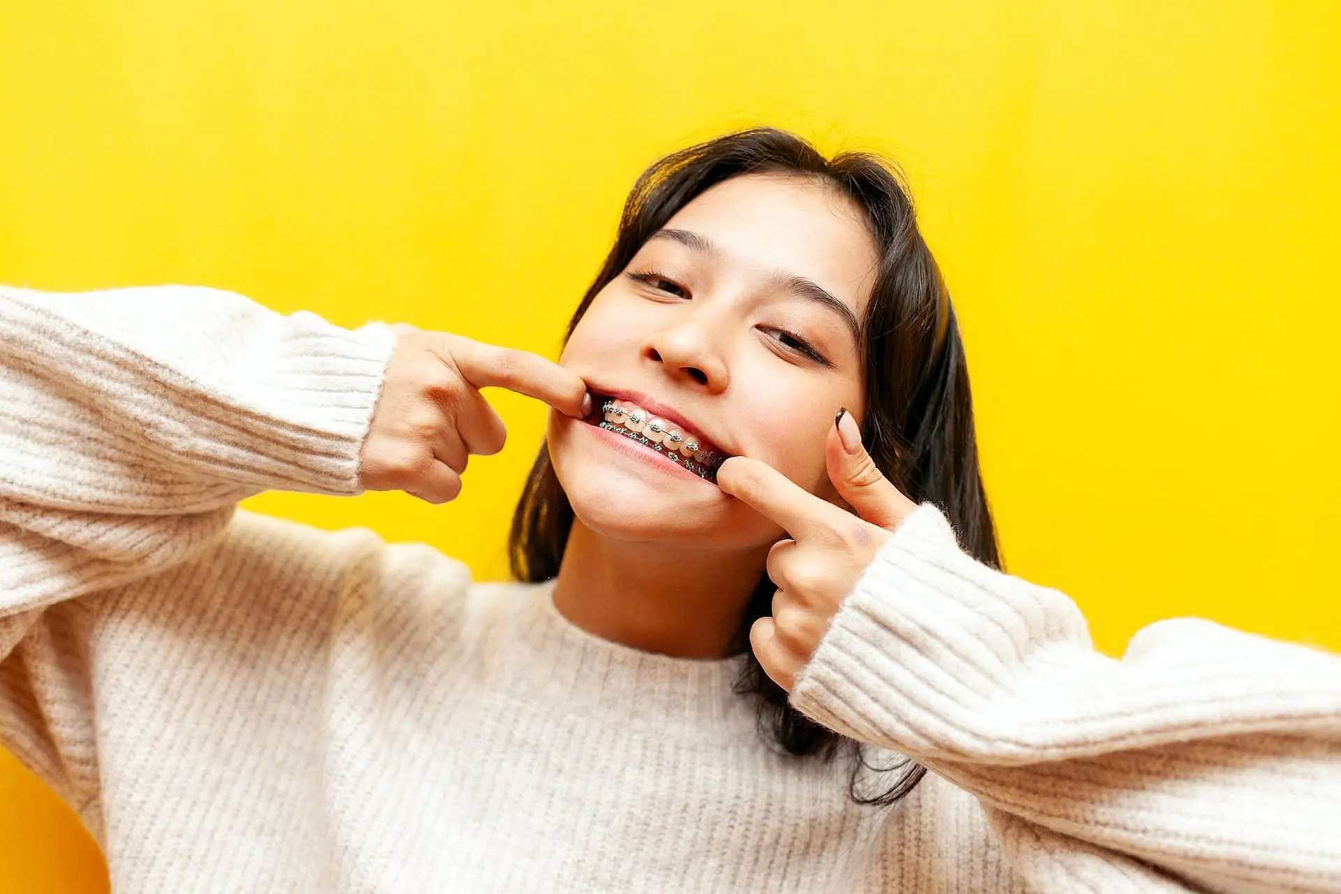 Showcasing the benefits of adult orthodontics, a person against a bright yellow background pulls back their lips to reveal braces—an example of what Orthodontic Design Co. Orthodontics offers in Madison and Huntsville, AL.