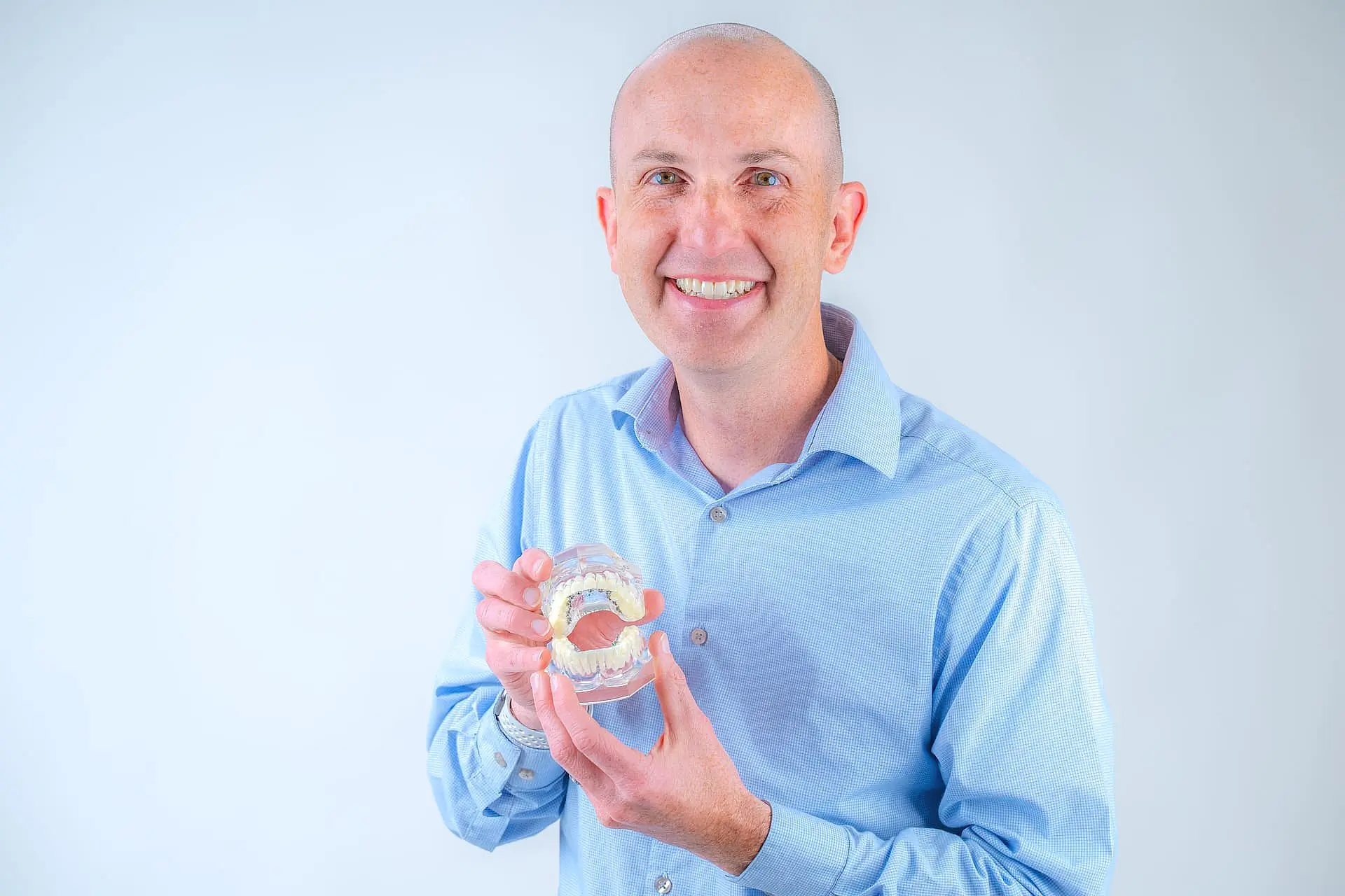 Dr. Adam Reynolds, a smiling man in a light blue shirt, holds a dental model showcasing the benefits of Adult Orthodontics at Orthodontic Design Co. Orthodontics in Madison and Huntsville, AL.