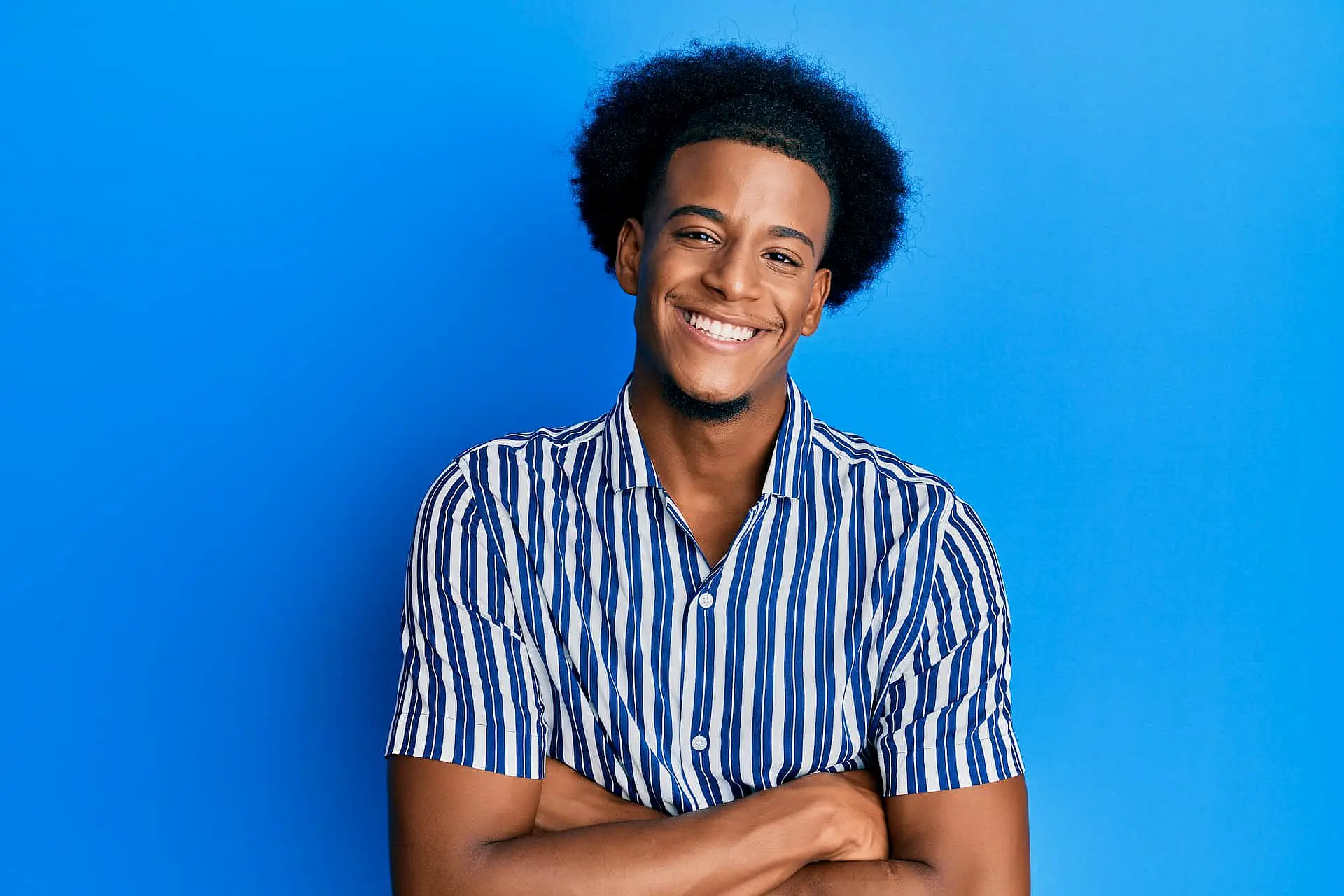 A smiling person with short curly hair, dressed in a blue and white striped shirt, stands confidently against a solid blue background—reflecting the results of adult orthodontics at Orthodontic Design Co. Orthodontics in Madison and Huntsville, AL.