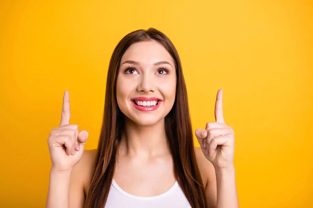 A smiling woman with long brown hair points upward with both index fingers against a solid yellow background, Schedule your free orthodontic consultation with Orthodontic Design Co. Orthodontics in Madison and Huntsville, AL!.