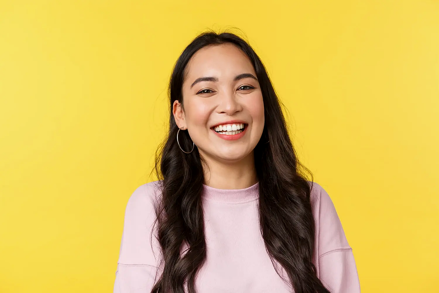 A woman with long dark hair and hoop earrings smiles in front of a yellow background for Orthodontic Design Co. and Best Orthodontist in Madison and Huntsville, AL.