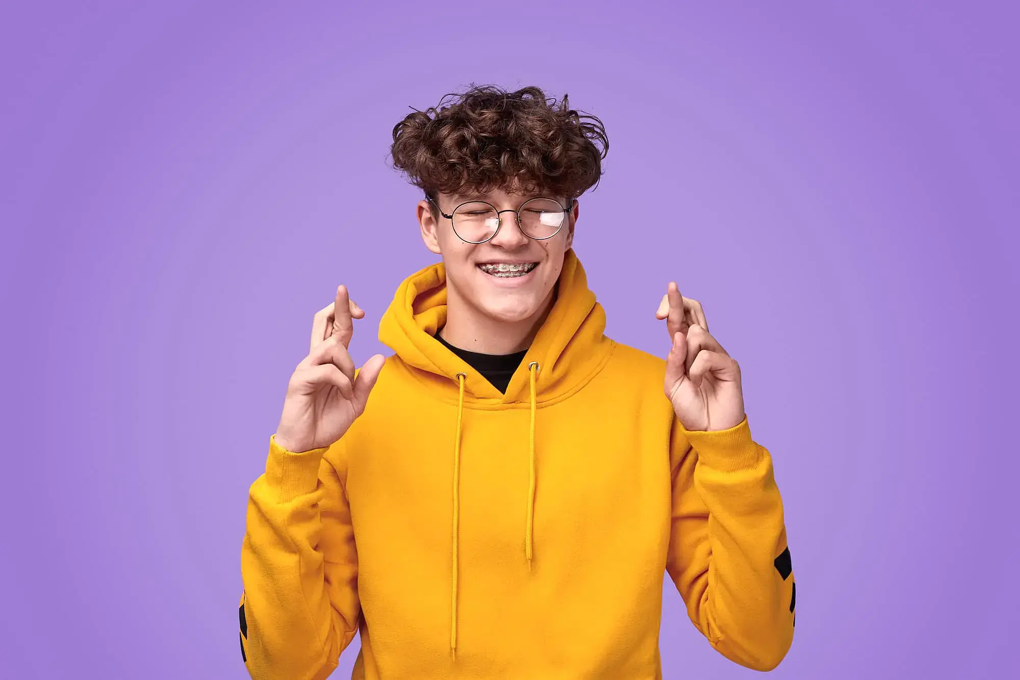 Smiling teen with curly hair and glasses in yellow hoodie crosses fingers, promoting Orthodontic Design Co. Orthodontics in Madison and Huntsville, AL, against a purple background. Smiling teen with curly hair and glasses in yellow hoodie crosses fingers, promoting Orthodontic Design Co. Orthodontics in Madison and Huntsville, AL, against a purple background.
