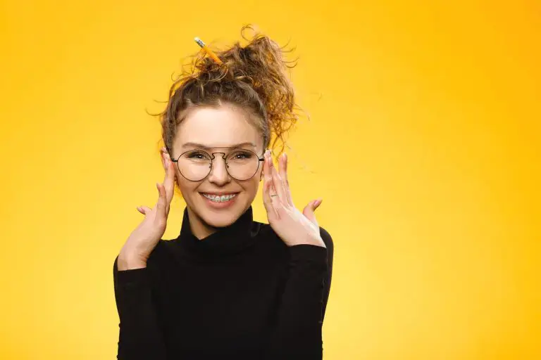 Smiling woman with glasses and braces, black turtleneck, pencil in curly hair, yellow backdrop for Orthodontic Design Co. Orthodontics in Madison and Huntsville, AL. Smiling woman with glasses and braces, black turtleneck, pencil in curly hair, yellow backdrop for Orthodontic Design Co. Orthodontics in Madison and Huntsville, AL.
