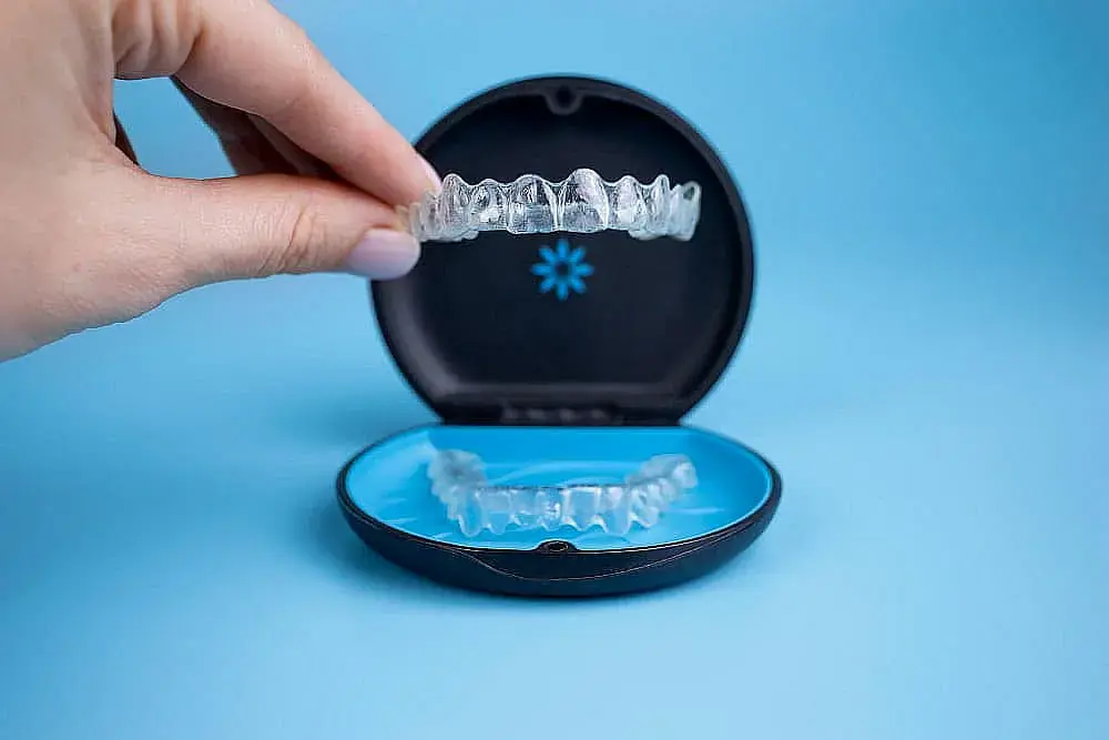 A hand holds a clear dental aligner over an open black and blue case with another aligner represent Invisalign payment at Orthodontic Design Co. Orthodontics in Madison and Huntsville, AL, on a blue background. A hand holds a clear dental aligner over an open black and blue case with another aligner represent Invisalign payment at Orthodontic Design Co. Orthodontics in Madison and Huntsville, AL, on a blue background.