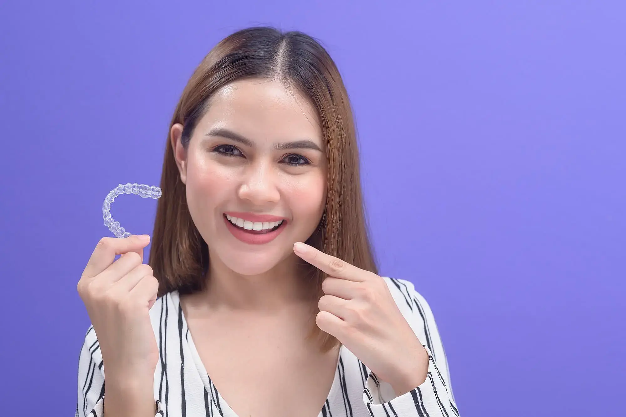 A woman smiles, holding a clear dental aligner and pointing to her teeth, representing Invisalign payment at Orthodontic Design Co. Orthodontics in Madison and Huntsville, AL, with a plain purple background. A woman smiles, holding a clear dental aligner and pointing to her teeth, representing Invisalign payment at Orthodontic Design Co. Orthodontics in Madison and Huntsville, AL, with a plain purple background.