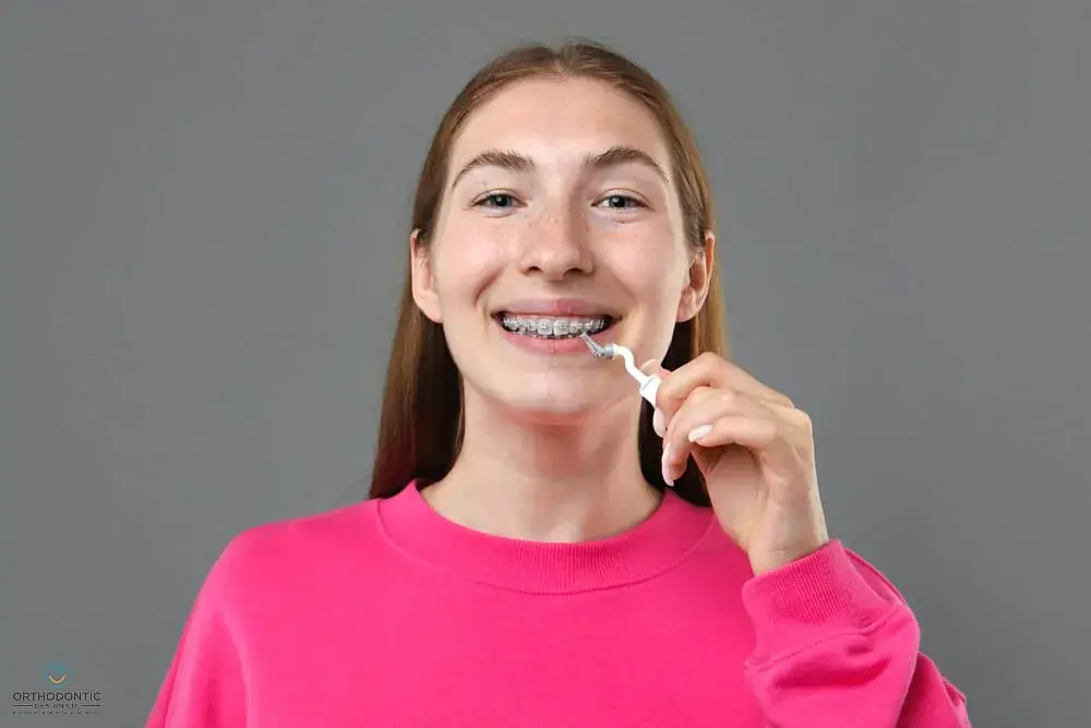 Smiling woman in pink sweatshirt uses a floss threader for braces at Orthodontic Design Co. Orthodontics in Madison and Huntsville, AL - How to Floss with Braces in Madison and Huntsville, AL. Smiling woman in pink sweatshirt uses a floss threader for braces at Orthodontic Design Co. Orthodontics in Madison and Huntsville, AL - How to Floss with Braces in Madison and Huntsville, AL.